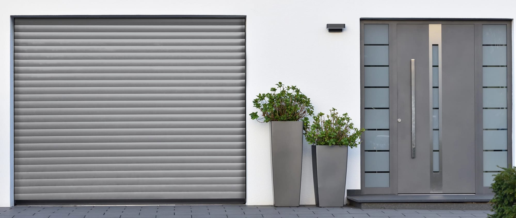 Façade d'une maison blanche avec une porte, deux pots de fleurs et une porte de garage enroulable Dooralux grise