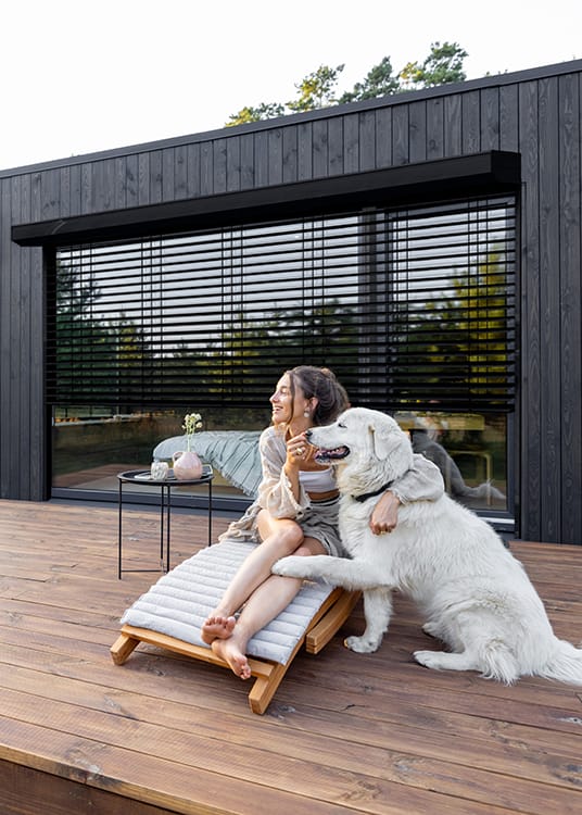 Vue depuis une terrasse en bois d'une femme et d'un chien posant devant des brise soleil orientables rénovation noir