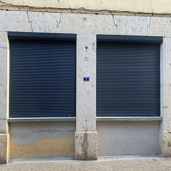 Vue extérieur depuis la rue de deux volets roulants renolux astrolux gris foncés sur une façade en pierre beige de la marque Flip
